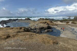 Glass Beach Fort Bragg Rockhounding: Wide view of the rugged coastline with rocky outcrops, tidal pools, and a few people searching for sea glass on the beach under a partly cloudy sky.