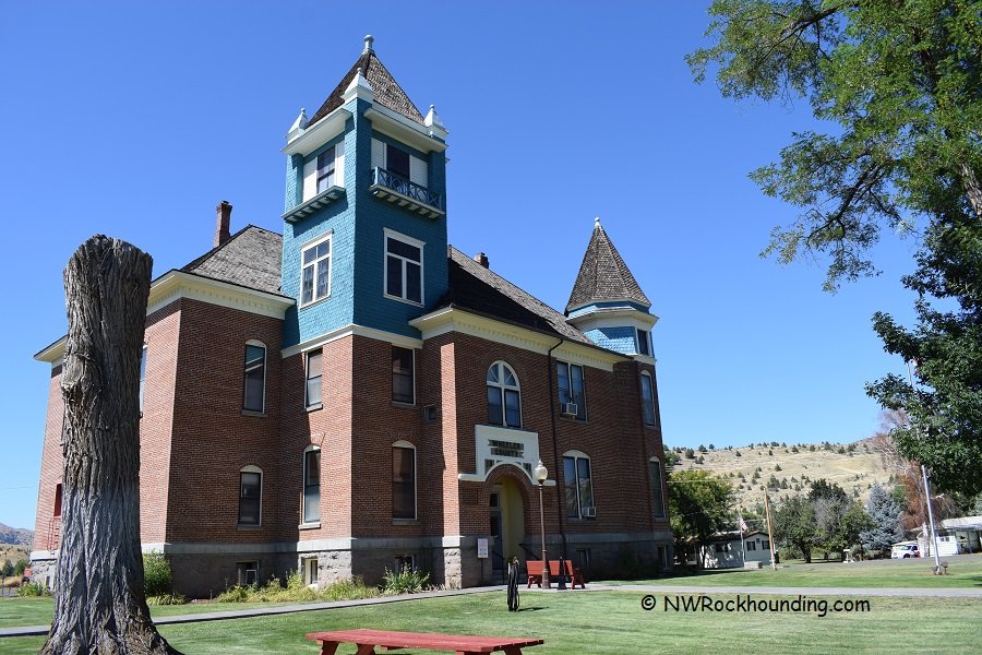 Wheeler High School Fossil Beds Rockhounding: The picture "Wheeler High School Fossil Beds Rockhounding historic school building" features a large, historic brick school building with blue and cream trim, two tall towers, and arched windows, set on a green lawn with a tree stump in the foreground.