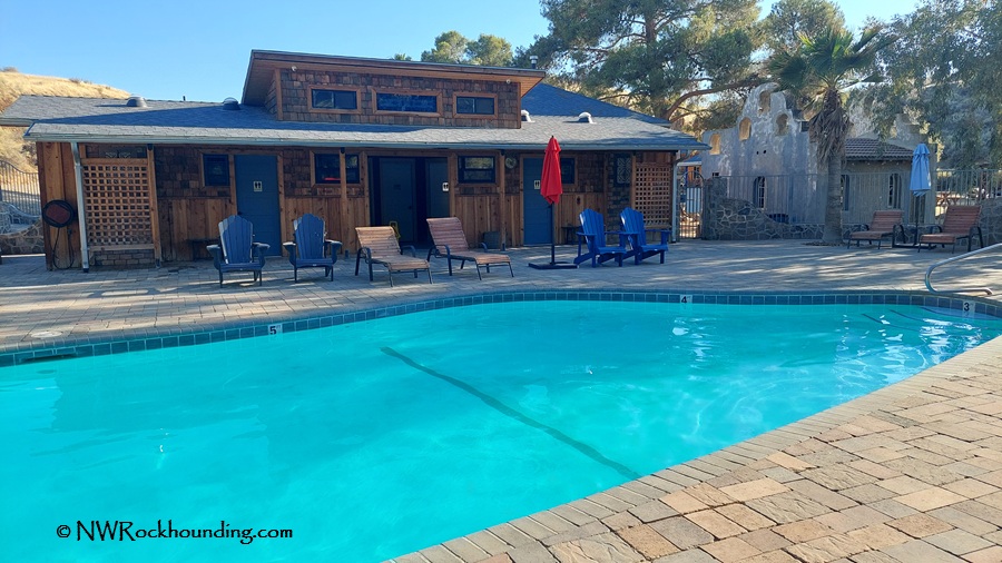 Panoche Hills Rockhounding in California near Mercey Hot Springs: The picture "Panoche Hills Rockhounding in California near Mercey Hot Springs pool and bathhouse" shows a bright blue swimming pool in front of a rustic wooden bathhouse with lounge chairs and umbrellas, surrounded by trees and rolling hills.