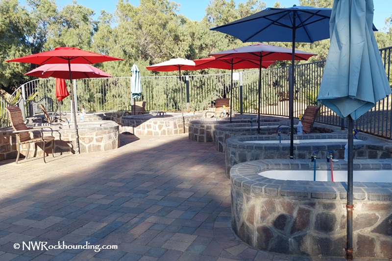 Panoche Hills Rockhounding in California near Mercey Hot Springs: The picture "Panoche Hills Rockhounding in California near Mercey Hot Springs stone soaking tubs" features multiple round stone soaking tubs with colorful umbrellas and lounge chairs, all set on a paved patio with trees and a metal fence in the background.