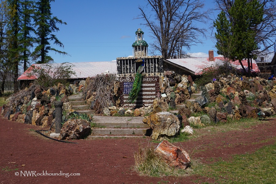 Petersen Rock Garden in Oregon: The picture "Petersen Rock Garden in Oregon American flag monument" features a rock and shell monument shaped like an American flag with the words 'God Bless America,' surrounded by rock walls, peacocks, and a grassy yard with trees and a house in the background.