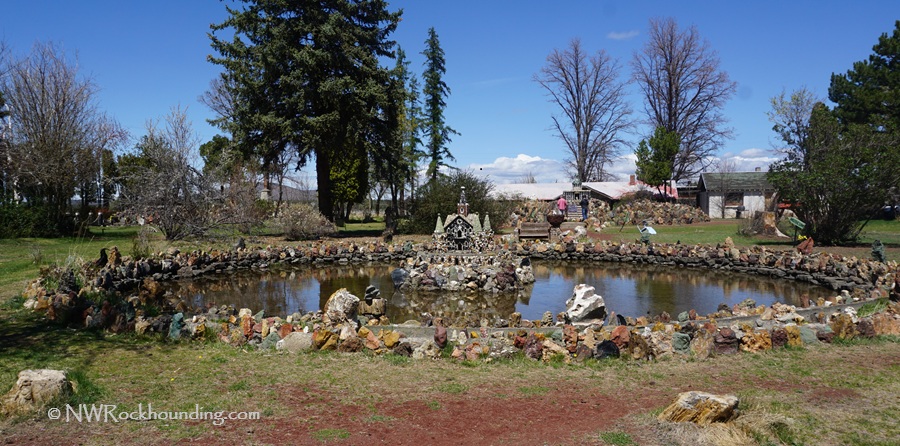 Petersen Rock Garden in Oregon: The picture "Petersen Rock Garden in Oregon round pond with rock border" features a round pond edged with colorful rocks, with miniature stone structures in the center and tall trees and grassy lawns in the background.
