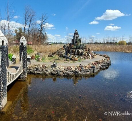 Petersen Rock Garden in Oregon: The picture "Petersen Rock Garden in Oregon castle island and bridge" shows a small stone footbridge leading to an island in a pond, featuring a miniature rock castle surrounded by water and tall grasses, under a blue sky with scattered clouds.