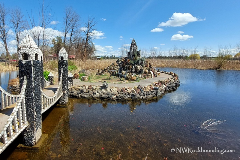 Petersen Rock Garden in Oregon: The picture "Petersen Rock Garden in Oregon castle island and bridge" shows a small stone footbridge leading to an island in a pond, featuring a miniature rock castle surrounded by water and tall grasses, under a blue sky with scattered clouds.