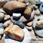 Selway River Rockhounding in Idaho: The picture "Selway River Rockhounding in Idaho – riverbank rocks" shows a close-up view of a collection of smooth, multicolored river rocks and pebbles scattered across a sandy riverbank.