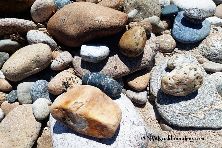 Selway River Rockhounding in Idaho: The picture "Selway River Rockhounding in Idaho – riverbank rocks" shows a close-up view of a collection of smooth, multicolored river rocks and pebbles scattered across a sandy riverbank.