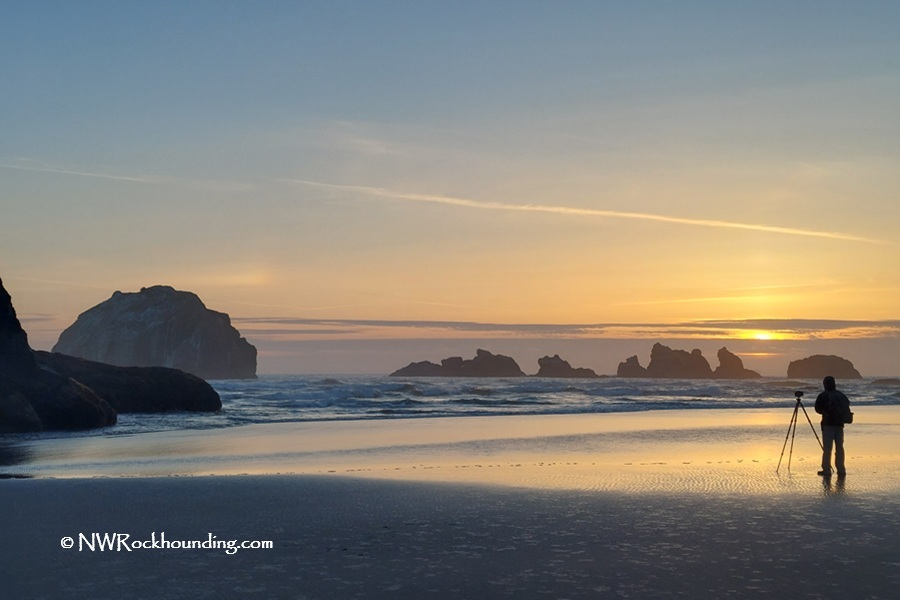 Bullard Beach Rockhounding - Treasures of Coquille River Mouth Gravel Bars in Oregon: The picture "Bullard Beach Rockhounding - Treasures of Coquille River Mouth Gravel Bars in Oregon – photographer at sunset with sea stacks" displays a person standing on the wet sand with a camera and tripod, capturing the colorful sunset over the ocean with dramatic sea stacks silhouetted on the horizon.