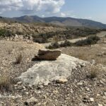 Gardiner Travertine in Montana: The picture "Gardiner Travertine in Montana – rock slab by dirt road" features a flat white slab of travertine with a chunk of rock resting on top, set on a rocky, arid hillside overlooking a dirt road and distant mountain ranges under a partly cloudy sky.