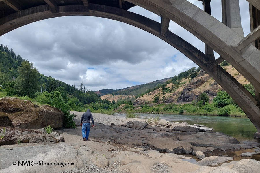 Myrtle Creek Rockhounding in Oregon: The picture "Myrtle Creek Rockhounding in Oregon – person exploring beneath arched bridge" shows a person in a gray hoodie and blue jeans walking on rocky ground beneath a large concrete arch bridge, with the river, distant hills, and cloudy sky stretching out in the background.