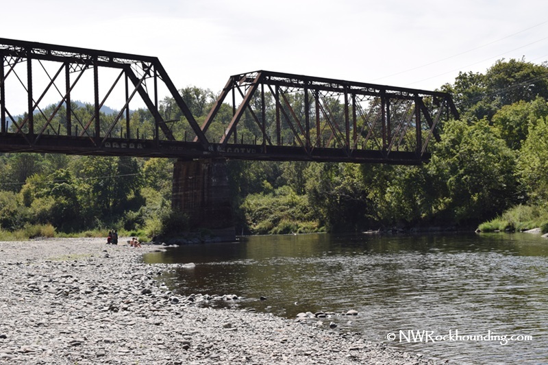 Myrtle Creek Rockhounding in Oregon: The picture "Myrtle Creek Rockhounding in Oregon – railroad bridge over rocky riverbank" features an old steel railroad bridge spanning a calm section of Myrtle Creek, with people visible relaxing on the large, rocky riverbank below, surrounded by lush green trees.