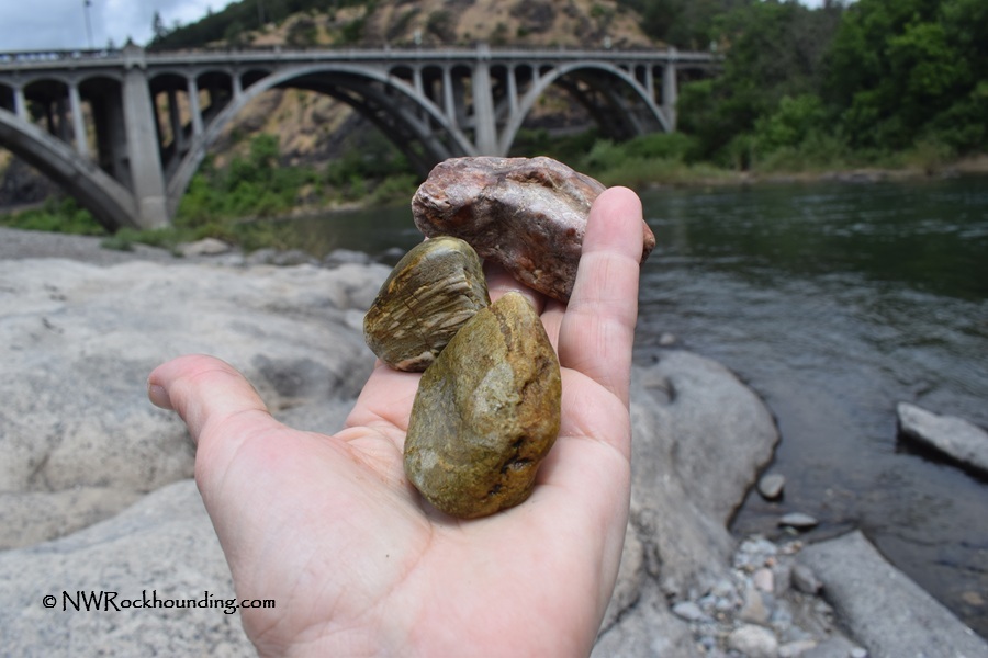 Myrtle Creek Rockhounding in Oregon: The picture "Myrtle Creek Rockhounding in Oregon – hand holding rocks by bridge" shows a person's hand holding three rough rocks, including greenish and reddish pieces, with the historic Myrtle Creek bridge and river in the background.