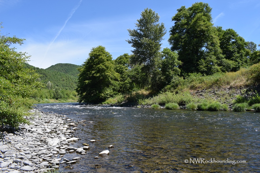 South Umpqua Rockhounding in Oregon: The picture "South Umpqua Rockhounding in Oregon – river with stony bank and trees" features a clear, flowing river bordered by a rocky shore and grasses on one side and a dense cluster of green trees beneath a blue sky.