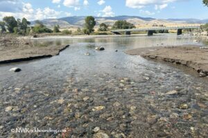 Yellowstone River Rockhounding in Montana: The picture "Yellowstone River Rockhounding in Montana – bridge and river scene" captures a calm section of the Yellowstone River with clear water revealing small rocks below, muddy banks on both sides, and a long bridge crossing the river with distant hills and blue sky in the background.