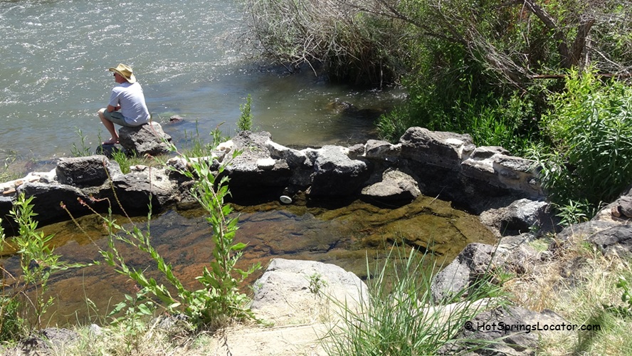 Bruneau River Canyon Rockhounding in Idaho: The picture "Bruneau River Canyon hot spring pool by river" features a natural hot spring pool edged with dark volcanic rocks, with a person in a hat sitting on the rocks beside a flowing river, surrounded by green bushes and grasses.