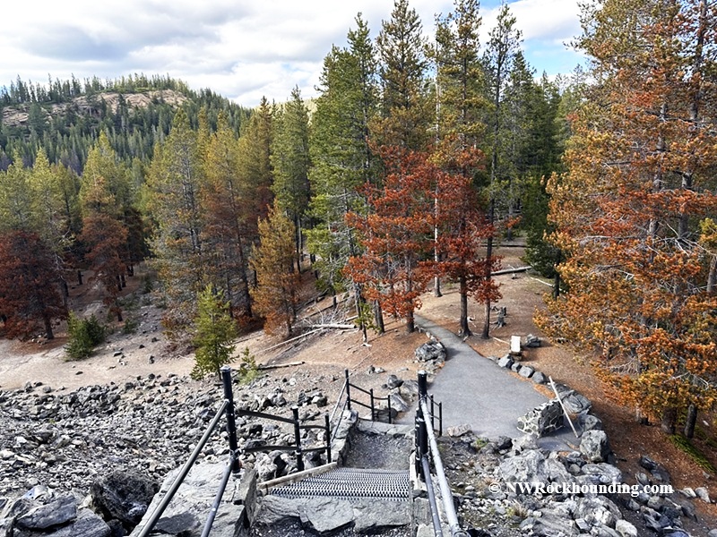 Big Obsidian Flow Oregon – Big Obsidian Flow Trailhead Big Obsidian Flow Oregon: The picture "Big Obsidian Flow Trailhead" shows a view down a metal stairway surrounded by black volcanic rocks, leading into a forested area with tall green and reddish trees and a paved trail below.