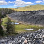 Big Obsidian Flow Oregon – Big Obsidian Flow Edge Big Obsidian Flow Oregon: The picture "Big Obsidian Flow Edge" captures the sharp, uneven edge of a massive obsidian lava flow rising above a grassy, marshy clearing and surrounded by evergreen forested hills.