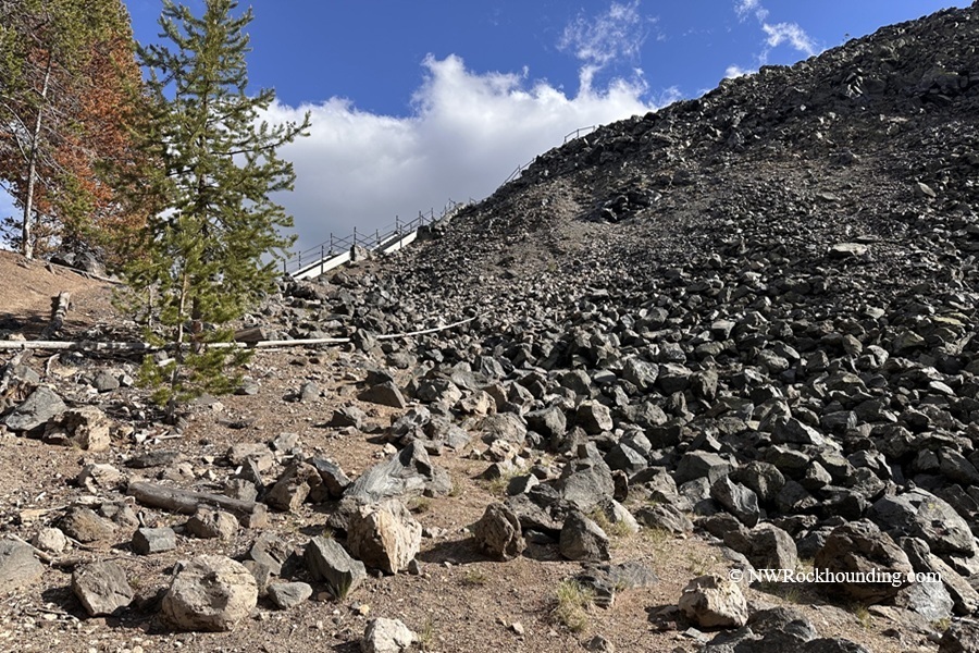Big Obsidian Flow Oregon – Big Obsidian Flow Slope Big Obsidian Flow Oregon: The picture "Big Obsidian Flow Slope" features a rocky volcanic hillside covered in dark angular stones, with scattered trees and a trail with railings visible at the top under a blue sky with clouds.